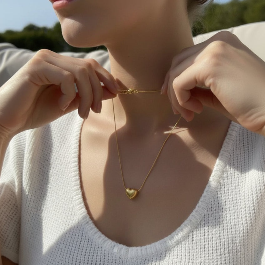 Person wearing a gold necklace with a heart pendant, set against a blurred natural background.